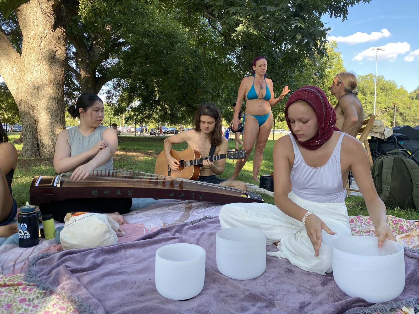 Members playing guzheng and guitar together in a park, with singing bowls set out on a blanket.